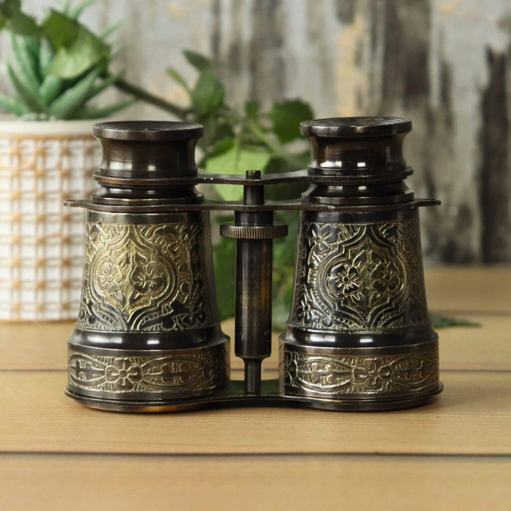 Front view of antique brass binoculars with intricate floral engravings and a black antique finish, standing on a wooden table.