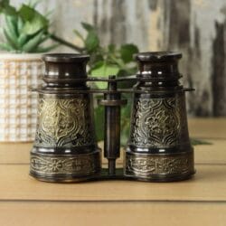 Front view of antique brass binoculars with intricate floral engravings and a black antique finish, standing on a wooden table.