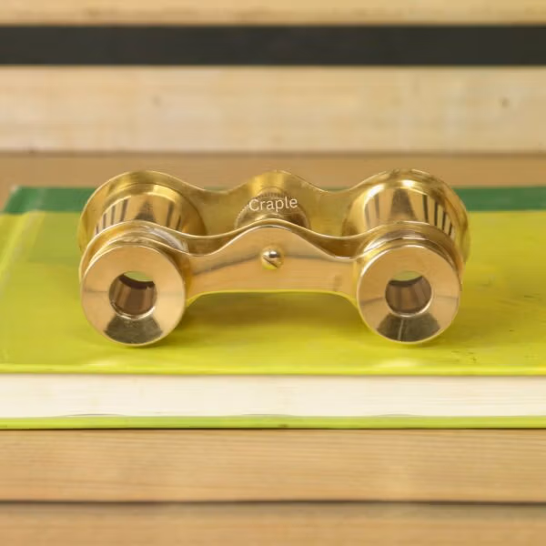 Top-down view of vintage brass opera glasses on a book, showing the bridge, knurled focus knob, and eyepiece design.
