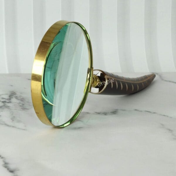 Angled side view of a large brass magnifying glass with a uniquely carved feather-shaped handle, resting on a marble surface.