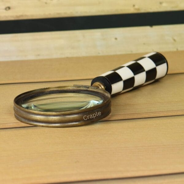 Angled view of a magnifying glass with an antique brass frame and a black and white checkerboard handle on a wood surface.