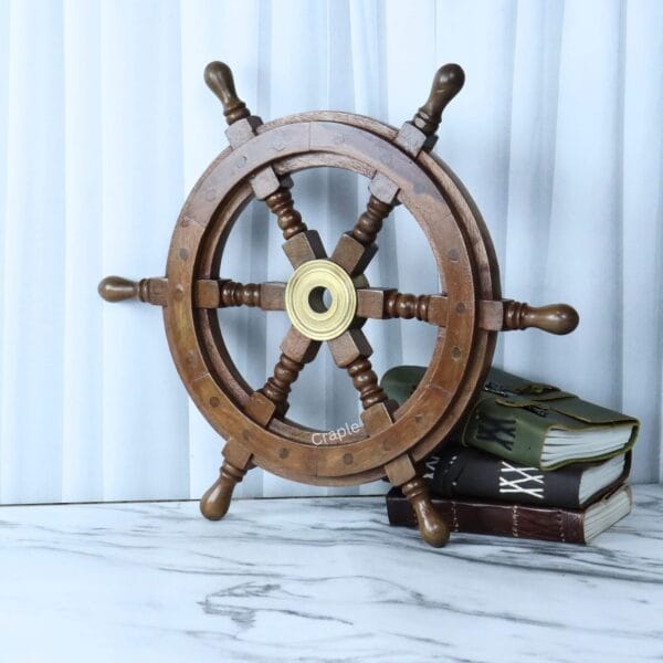 A dark wood decorative pirate ship helm with a brass hub, shown leaning next to a stack of vintage books.