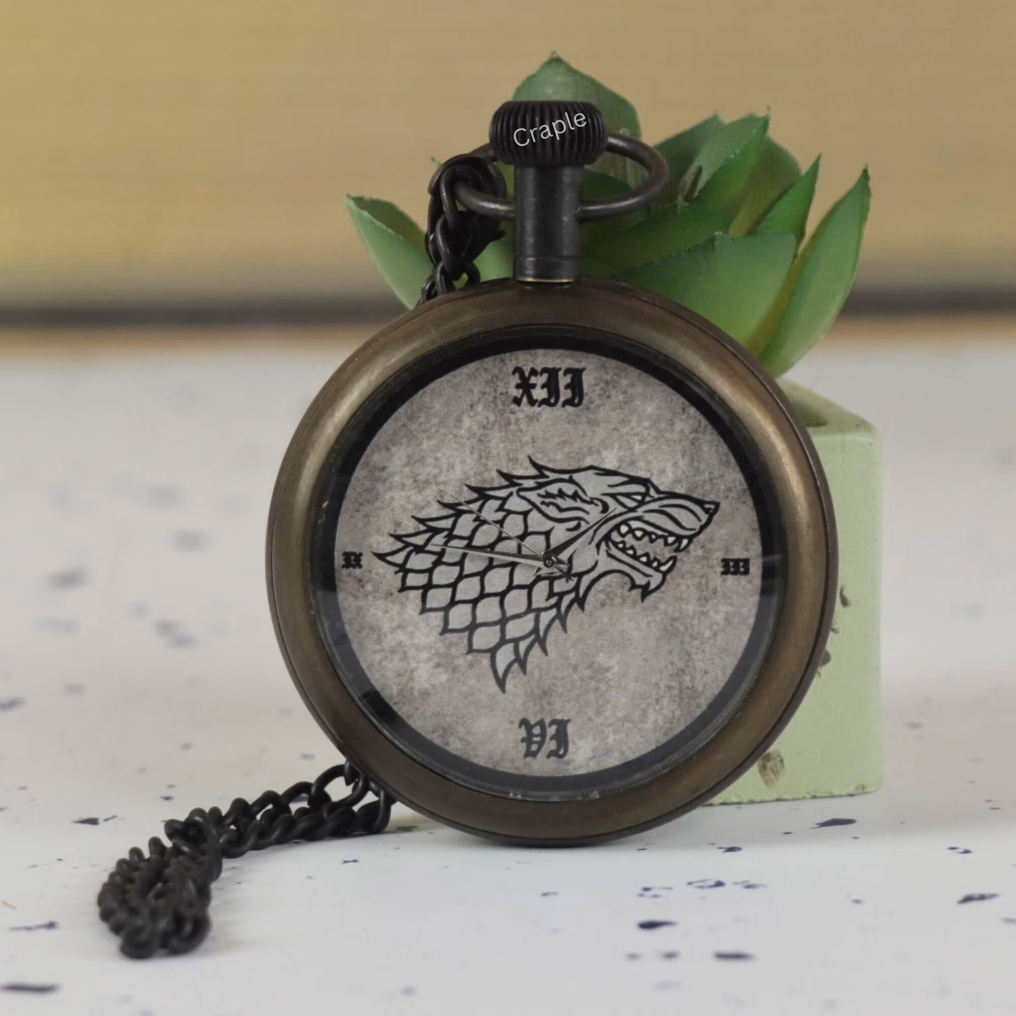An antique brass pocket watch featuring a Direwolf sigil on the dial, propped against a small succulent.