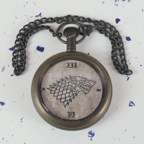 The Direwolf sigil pocket watch lying on a flat surface with its dark antique chain spread out above it.