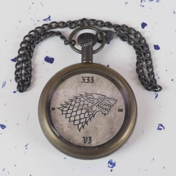The Direwolf sigil pocket watch lying on a flat surface with its dark antique chain spread out above it.