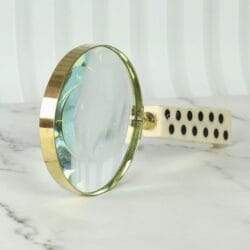 A decorative magnifying glass with a unique domino-style handle and a shiny brass rim, resting on a marble surface.
