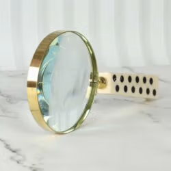 A decorative magnifying glass with a unique domino-style handle and a shiny brass rim, resting on a marble surface.