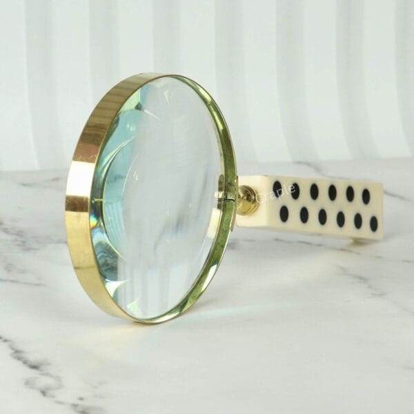 A decorative magnifying glass with a unique domino-style handle and a shiny brass rim, resting on a marble surface.