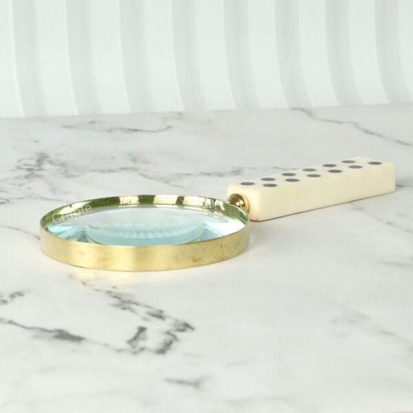 A 5X reading magnifier with a domino handle and polished brass frame, shown lying flat on a marble-textured surface.
