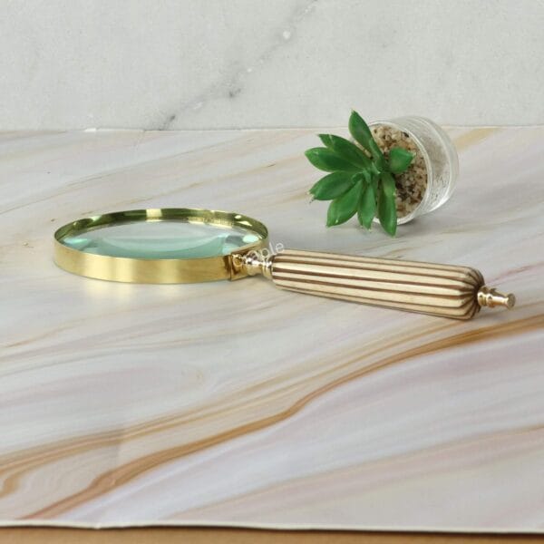 A decorative brass magnifying glass with a fluted striped handle, resting on a marble desk next to a small succulent.