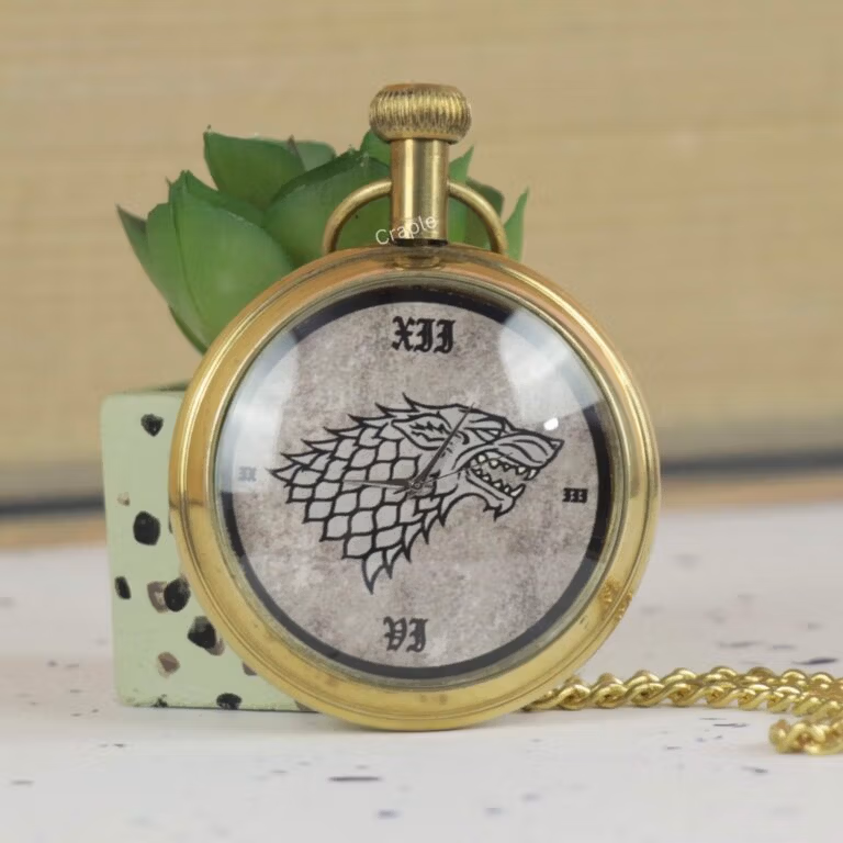 Vintage-style brass pocket watch featuring the Game of Thrones Direwolf sigil, resting against a decorative plant.