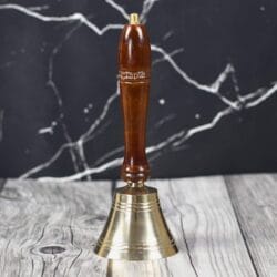 Handcrafted brass hand bell with a polished brown wooden handle, standing on a rustic wood surface.