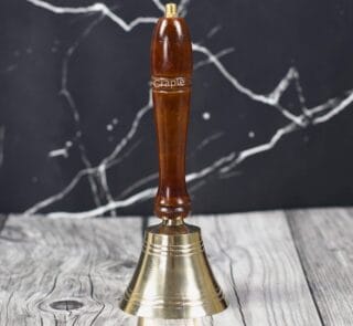 Handcrafted brass hand bell with a polished brown wooden handle, standing on a rustic wood surface.