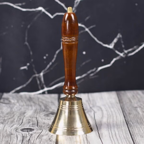 Handcrafted brass hand bell with a polished brown wooden handle, standing on a rustic wood surface.