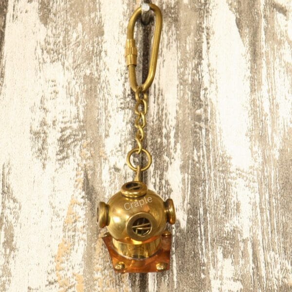 The nautical brass diver helmet keychain hanging by its carabiner clip against a rustic white wood background.