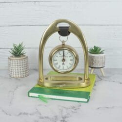 Golden aluminum hanging desk clock with a classic white face, suspended from an elegant arch stand on a marble tabletop.