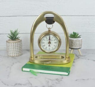 Golden aluminum hanging desk clock with a classic white face, suspended from an elegant arch stand on a marble tabletop.