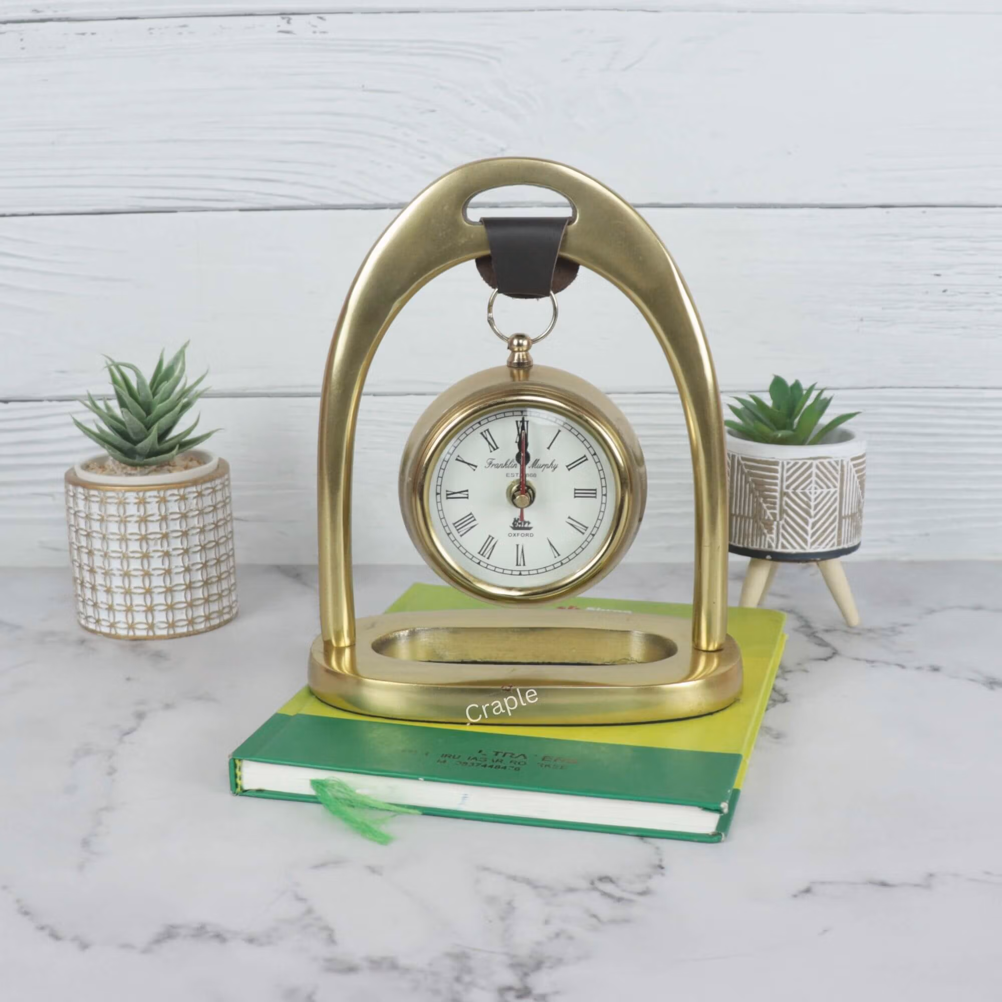 Golden aluminum hanging desk clock with a classic white face, suspended from an elegant arch stand on a marble tabletop.