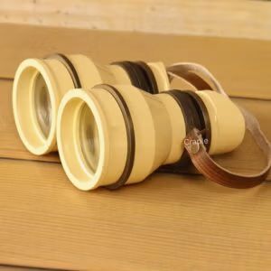 Retro light yellow aluminum binoculars with angled eyecups and leather strap on wooden table – vintage decorative binoculars for home decor or unique gift.