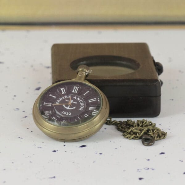 The Marine Anchor pocket watch and its brass chain displayed next to its closed dark wood presentation box.