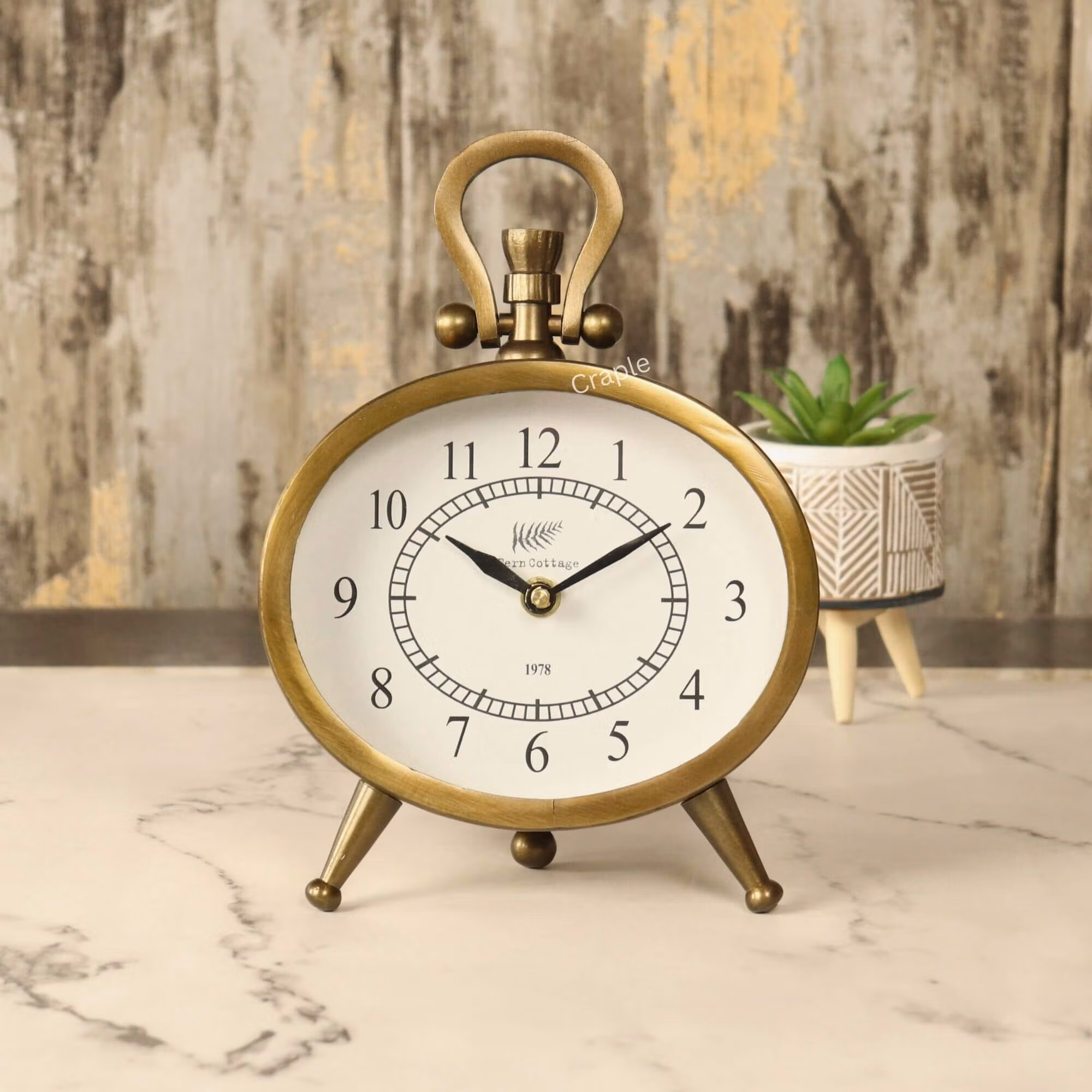 Oval Antique Aluminium Clock-1 Antique oval desk clock with an antique brass finish, featuring a white face and black numerals, standing on a marble tabletop.