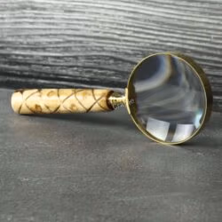 Vintage-style magnifying glass with a patterned wood handle and brass rim, standing upright on a dark surface.