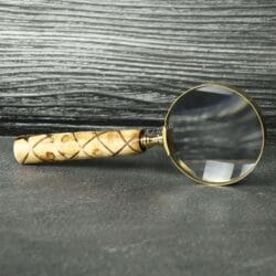 Handheld brass magnifying glass with a 5X lens and a patterned swirl wooden handle, resting on a slate tabletop.