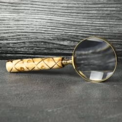 Handheld brass magnifying glass with a 5X lens and a patterned swirl wooden handle, resting on a slate tabletop.