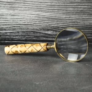 Handheld brass magnifying glass with a 5X lens and a patterned swirl wooden handle, resting on a slate tabletop.