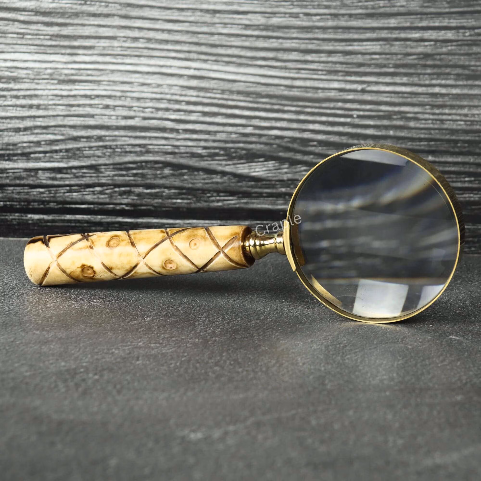 Patterned Swirl Handle Brass Magnifier-2 Handheld brass magnifying glass with a 5X lens and a patterned swirl wooden handle, resting on a slate tabletop.