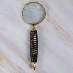 Vintage-style magnifying glass with a dark textured ribbed handle and a shiny brass-rimmed lens, shown on a marble surface.