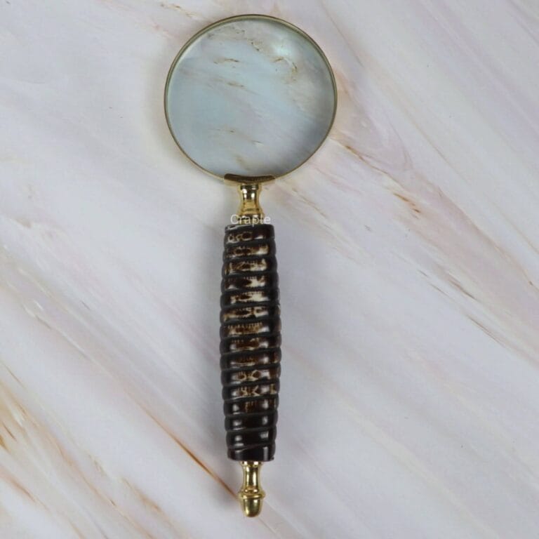Vintage-style magnifying glass with a dark textured ribbed handle and a shiny brass-rimmed lens, shown on a marble surface.