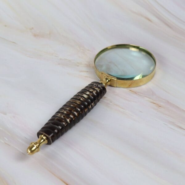 Angled view of the decorative brass magnifier, highlighting the texture and detail of the dark ribbed handle.