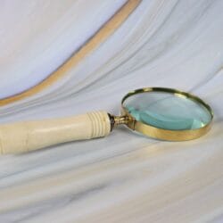 Side view of a vintage-style magnifying glass with a bone-style handle and shiny brass rim, resting on a marble surface.