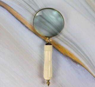 Vintage-style brass magnifying glass with a 5X lens and a carved, ergonomic bone-style handle against a marble background.