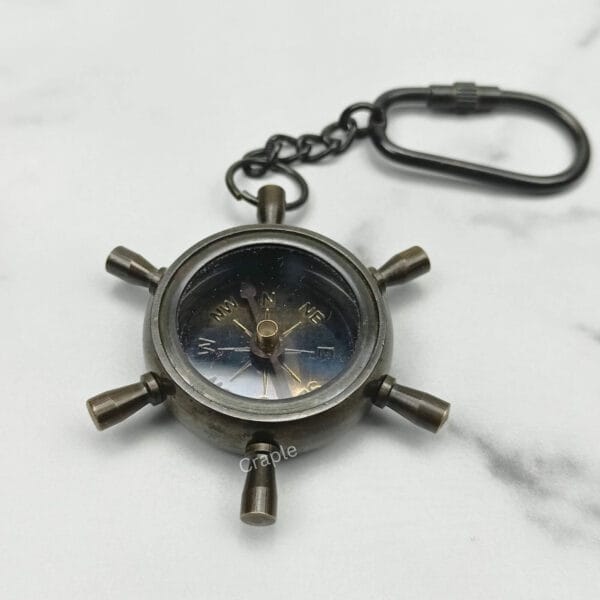 Top-down view of a nautical shipwheel compass keychain with a black antique finish on a marble surface.