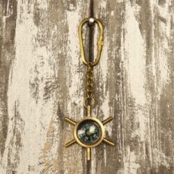 The nautical shipwheel compass keychain hanging from its brass carabiner clip against a rustic wood surface.