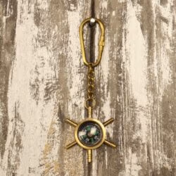 The nautical shipwheel compass keychain hanging from its brass carabiner clip against a rustic wood surface.