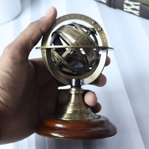 A hand holding the small brass armillary sphere to demonstrate its compact, desktop size.