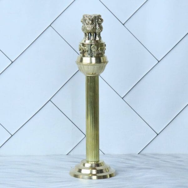 A full-length studio photograph of the tall brass Ashoka Pillar against a clean, white background.