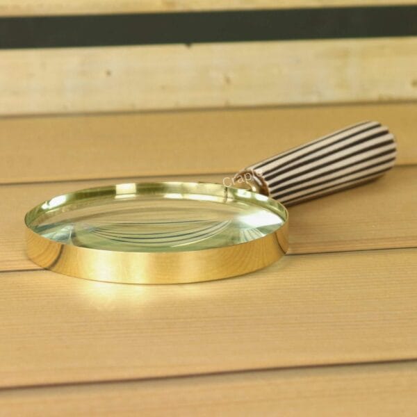 Handheld brass magnifying glass with a black and white striped handle, lying on a wooden desk.