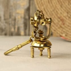 Front view of a miniature brass theodolite keychain designed as a vintage surveying instrument with a tripod base.