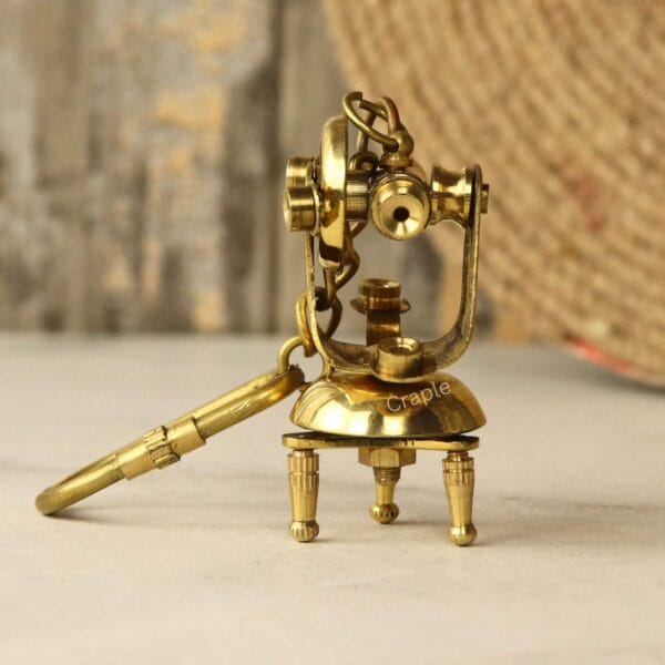 Front view of a miniature brass theodolite keychain designed as a vintage surveying instrument with a tripod base.