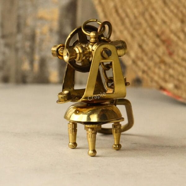 Angled side view of the shiny brass theodolite keychain, showing the intricate details of the miniature surveyor's tool.