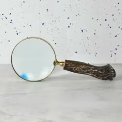 Vintage-style magnifying glass with a rustic tree trunk handle and a shiny brass frame, lying on a marble surface.