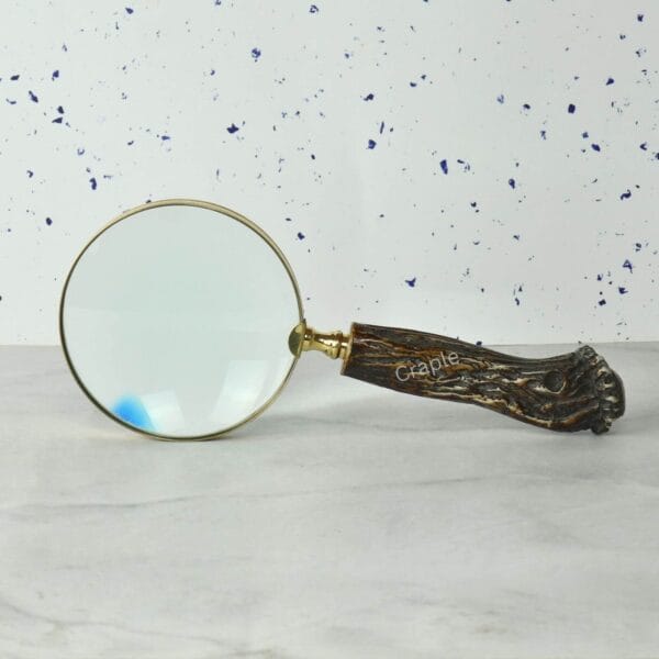Vintage-style magnifying glass with a rustic tree trunk handle and a shiny brass frame, lying on a marble surface.