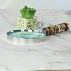 Handheld magnifier with a turned resin handle and nickel frame lying on a white marble desk next to a plant.