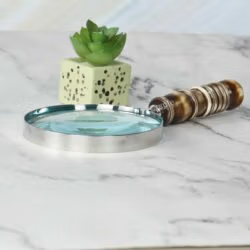 Handheld magnifier with a turned resin handle and nickel frame lying on a white marble desk next to a plant.