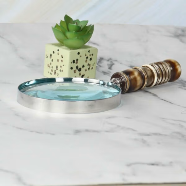 Handheld magnifier with a turned resin handle and nickel frame lying on a white marble desk next to a plant.
