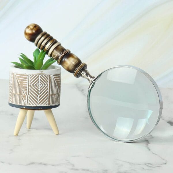 A decorative magnifying glass with a brown and white resin handle, propped against a stylish planter on a marble surface.